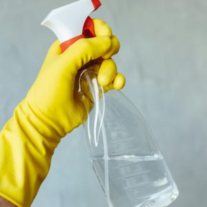Yellow gloved hand holding a transparent cleaning spray bottle against a neutral background.