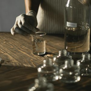 Crop anonymous female in latex gloves making fluid for perfume among glass jars on wooden table
