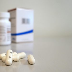 Close-up image of white capsules with medicine bottle and box in the background.