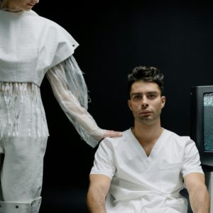 A man sits in white attire while a woman in futuristic clothing stands beside in a modern medical setting.